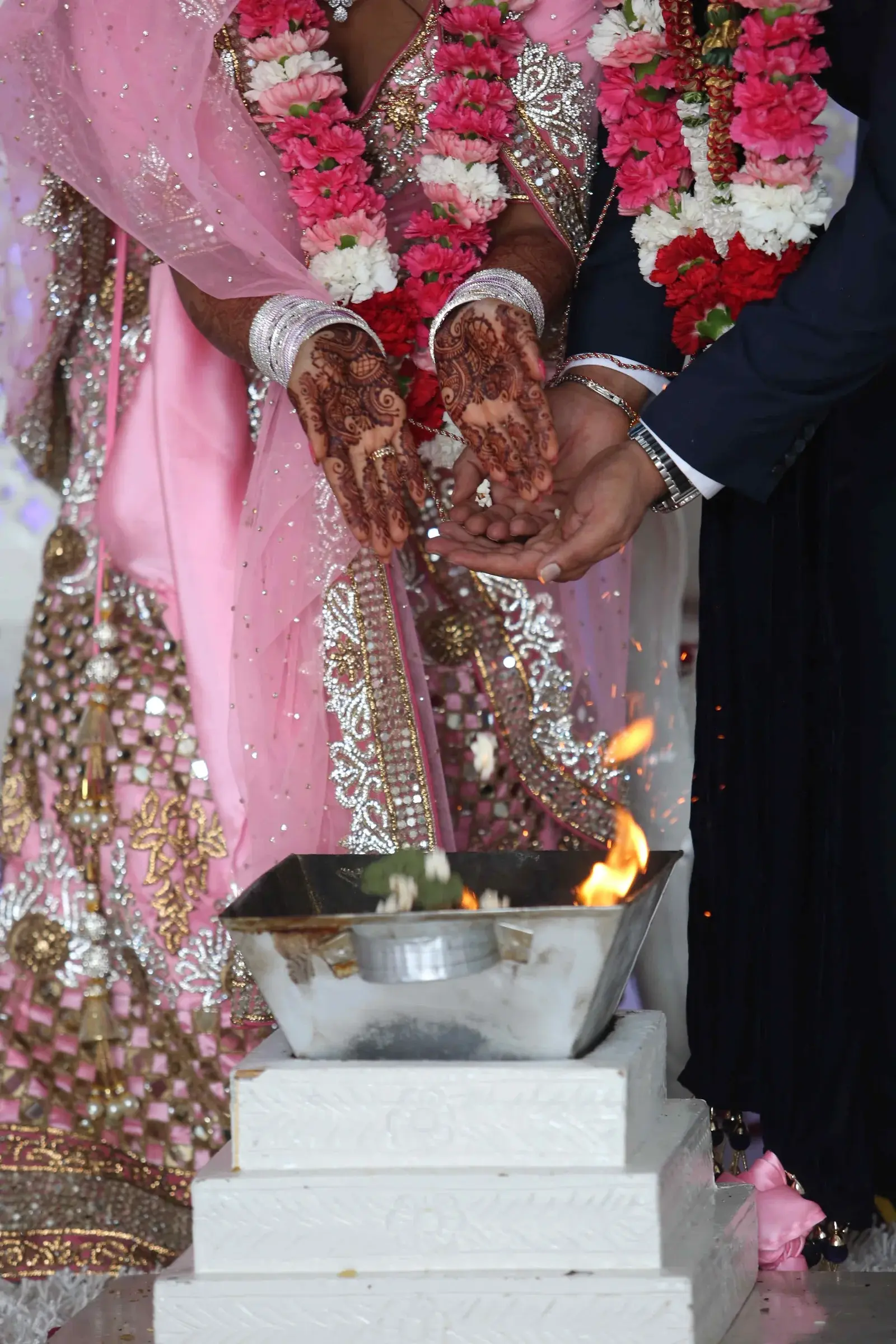 Bride in pink sari with detailed mehndi on hands extended over fire ritual vessel at Hindu wedding