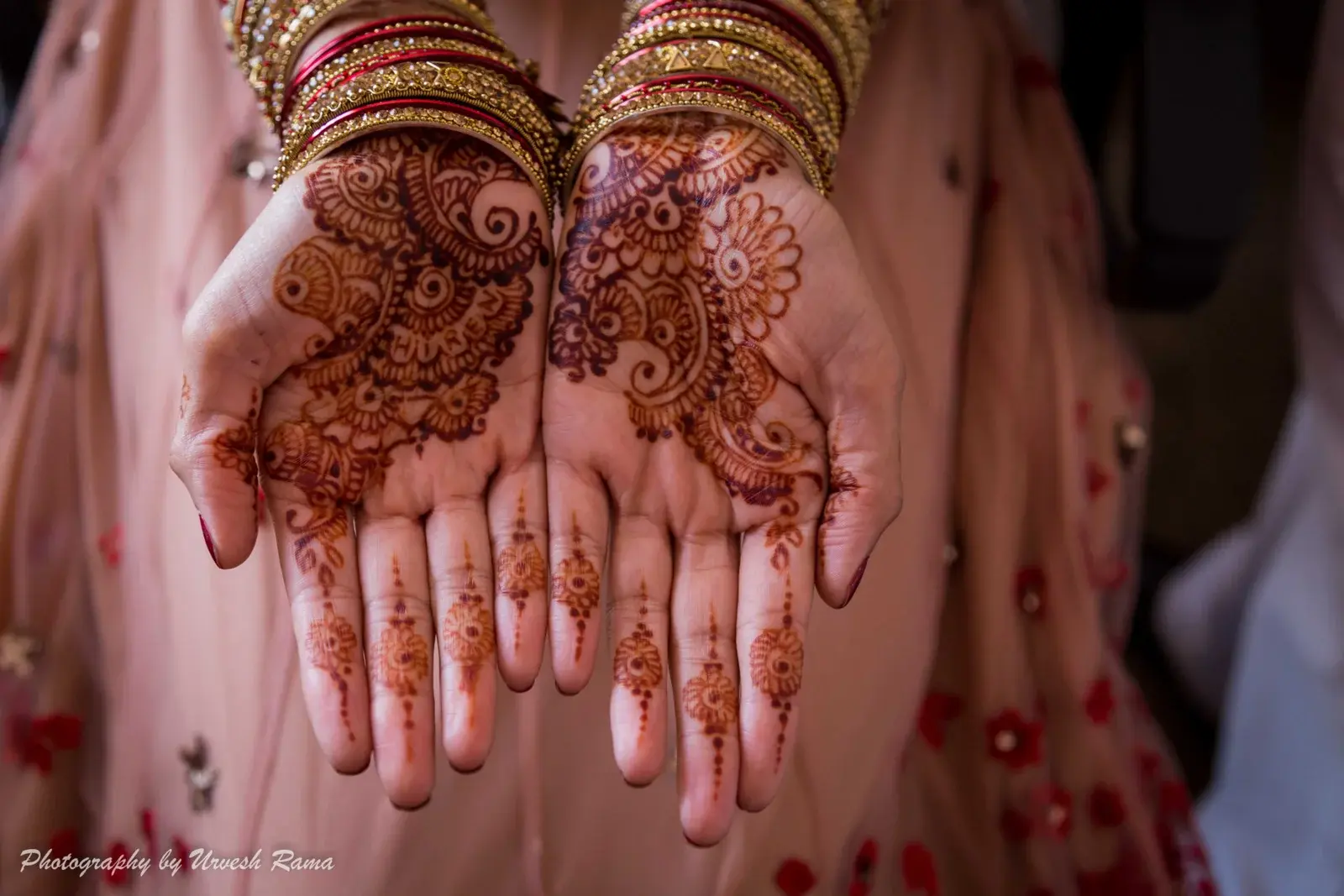 Bride displaying full-palm intricate bridal mehndi with gold bangles against pink floral lehenga, professional photography