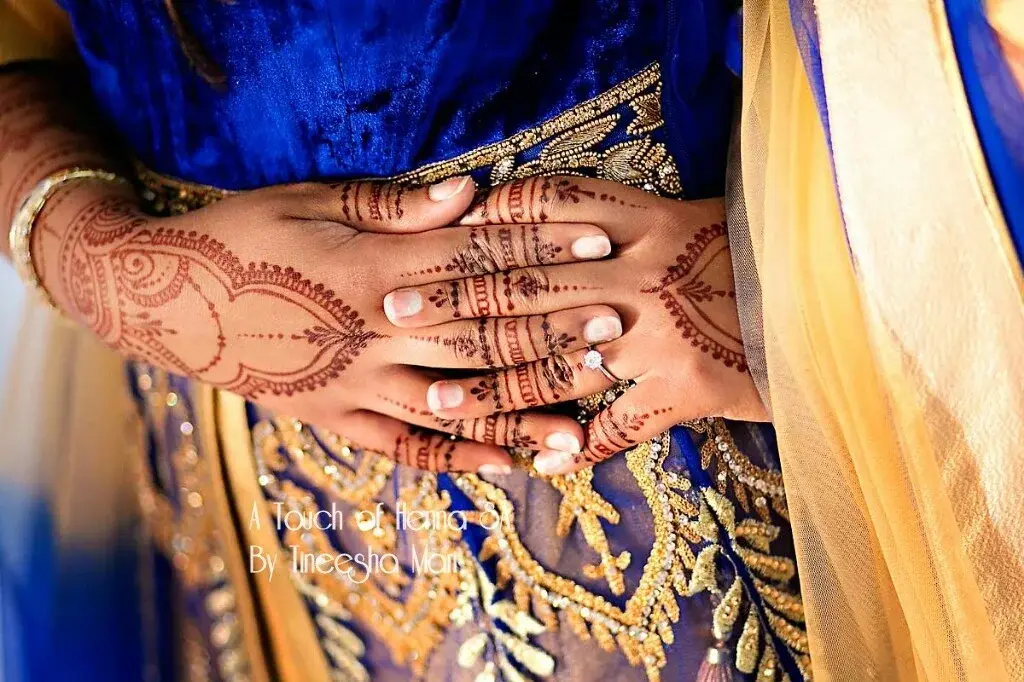 Bride in blue and gold lehenga displaying full-arm mehndi with diamond engagement ring, professional photography