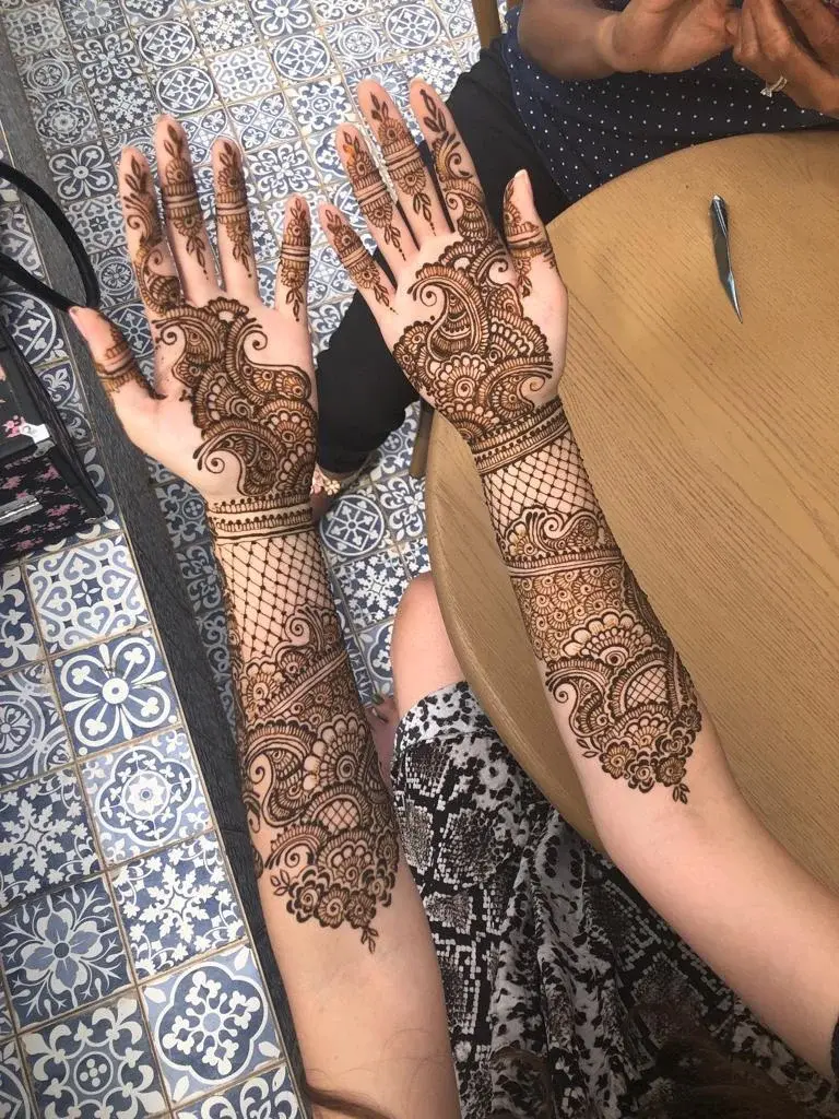 Full-arm bridal mehndi with intricate paisley and lattice covering both forearms and palms, applied at blue-tiled surface