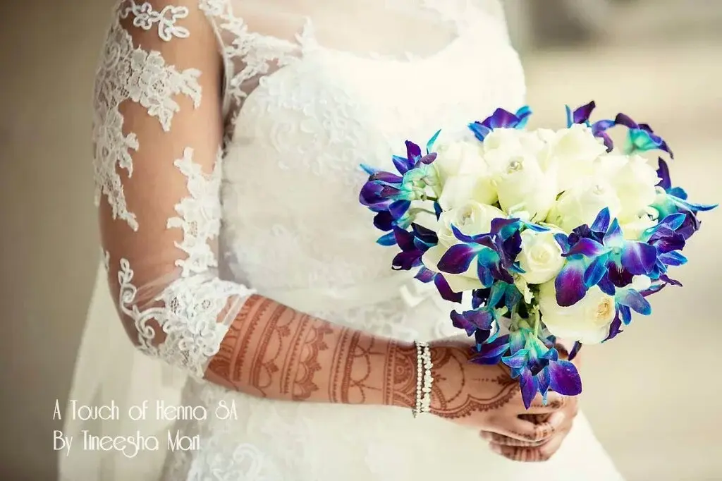 Bride in white wedding dress holding bouquet of blue orchids and white roses, mehndi visible on forearms