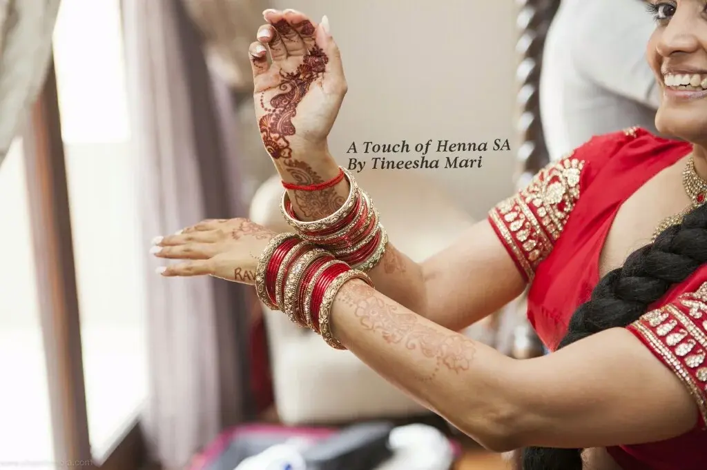 Bride Nalini in red bridal attire with gold bangles, smiling while having mehndi applied
