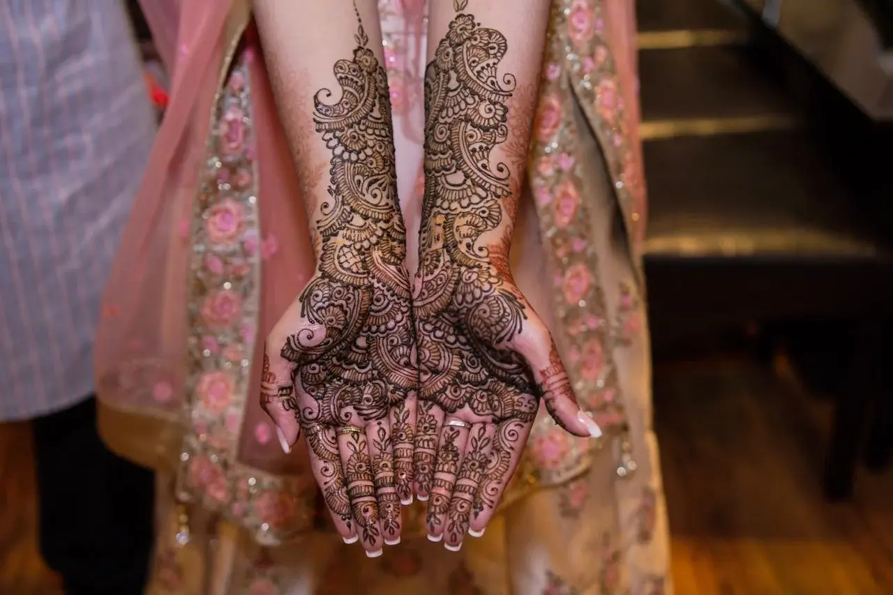 Full-arm bridal mehndi with sweeping floral and scroll design on both arms extended, bride in pink lehenga