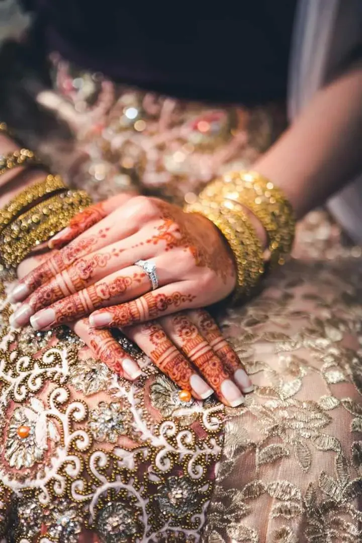 Two sets of mehndi-covered hands stacked together with gold bangles and diamond ring, ornate gold embroidered fabric