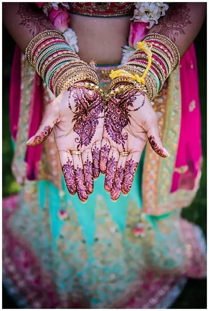 Bride displaying palm mehndi with gold bangles and floral garland, wearing teal and pink bridal lehenga