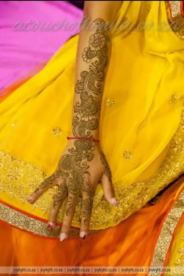Intricate full-arm mehndi in warm tones on hand and forearm against yellow bridal saree, professional photography