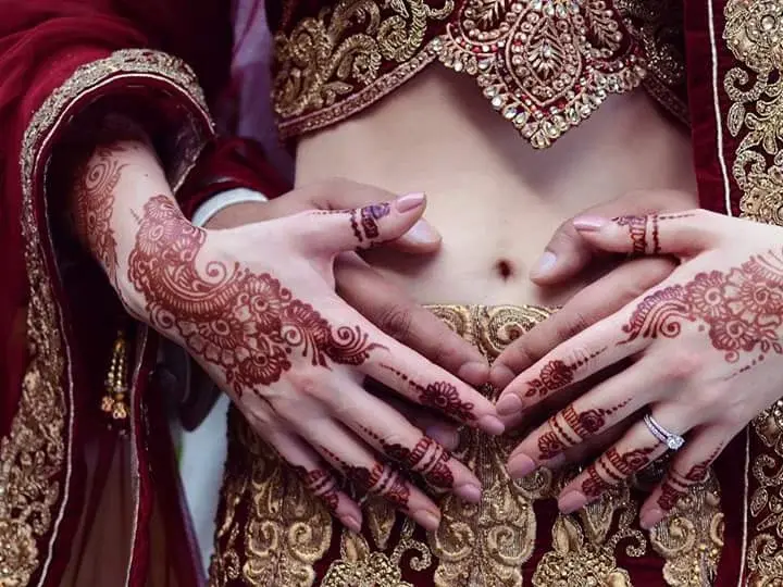 Bride in red and gold lehenga displaying intricate full-hand mehndi with floral detailing, diamond ring on finger